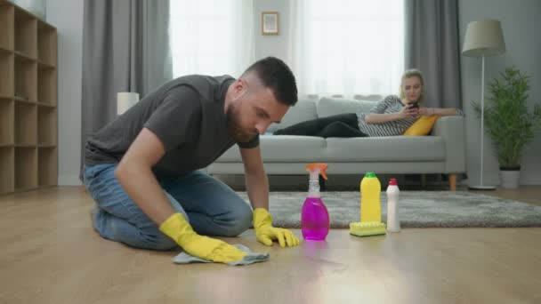 Homme fatigué nettoie parquet, et la femme se trouve sur le canapé avec smartphone — Video