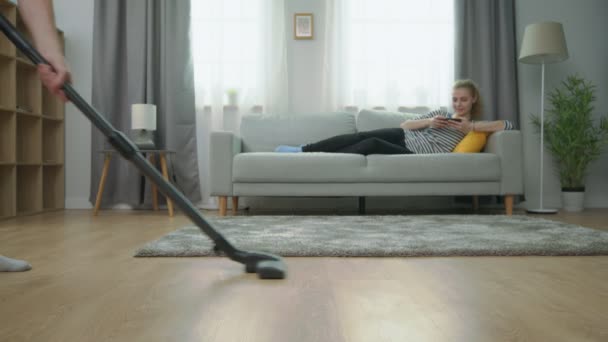 Suami lelah mengosongkan lantai parket, dan istrinya bermain di smartphone di sofa — Stok Video