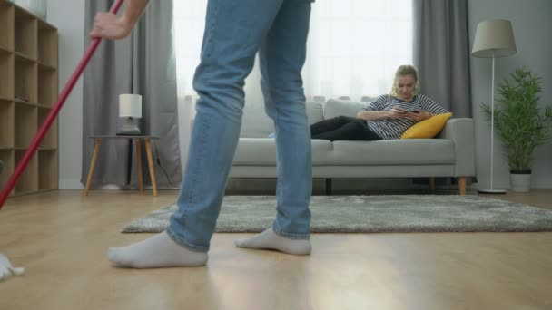 Tired man mopping parquet floor and his girlfriend play on smartphone on sofa — Stock Video