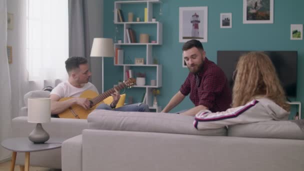 Panning di giovane suona la chitarra per i suoi amici in un accogliente soggiorno . — Video Stock
