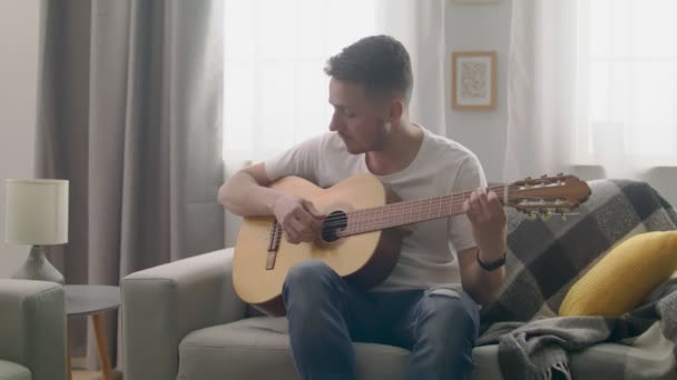 Handheld tiro de homem atraente toca guitarra em sua acolhedora sala de estar . — Vídeo de Stock