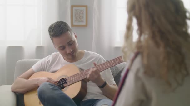 Handheld shot of man spelar gitarr för sina flickvänner i ett mysigt vardagsrum. — Stockvideo