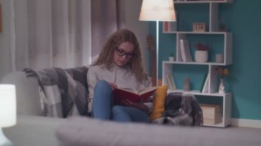 Genç ve güzel bir kadın akşamları oturma odasında kağıt kitap okuyor.