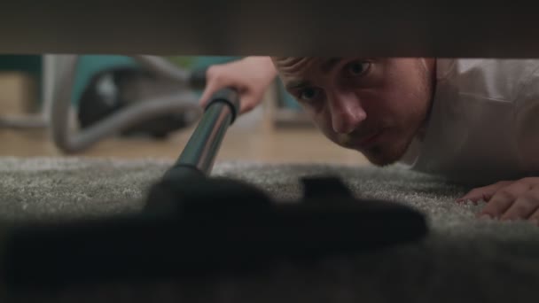 Young man cleaning a floor under a bed lying on the floor using vacuum cleaner — 图库视频影像