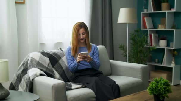 Mladá veselá dívka s chytrým telefonem ve svém útulném obývacím pokoji — Stock video