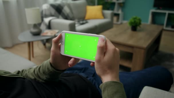 Homem em casa Sitng em um sofá usando Smartphone com tela de Mock-up verde — Vídeo de Stock