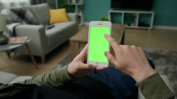 Чоловік вдома сидить на дивані за допомогою смартфона з зеленим макетним екраном — стокове відео