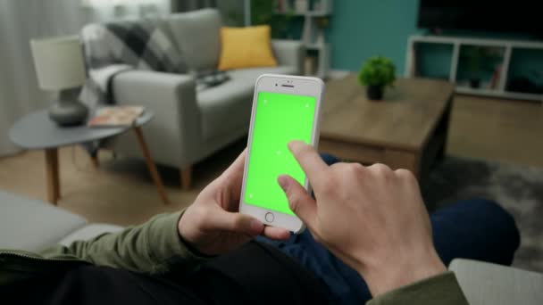 Чоловік вдома сидить на дивані за допомогою смартфона з зеленим макетним екраном — стокове відео