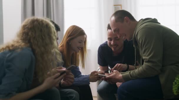 Feliz grupo de amigos usando teléfonos sentarse en el sofá interior riendo hablando divertirse — Vídeo de stock