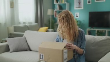 Genç bir kadın kanepede karton bir paket açıyor.