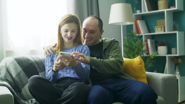 Ένα ζευγάρι μαζί χρησιμοποιώντας ένα smartphone στον καναπέ στο σαλόνι του — Αρχείο Βίντεο