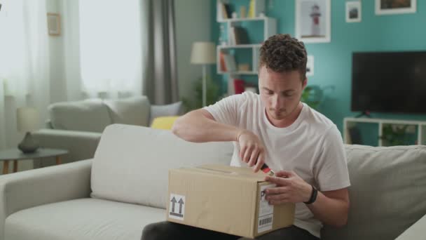 Jongeman in zijn woonkamer opent kartonnen doos pakket — Stockvideo