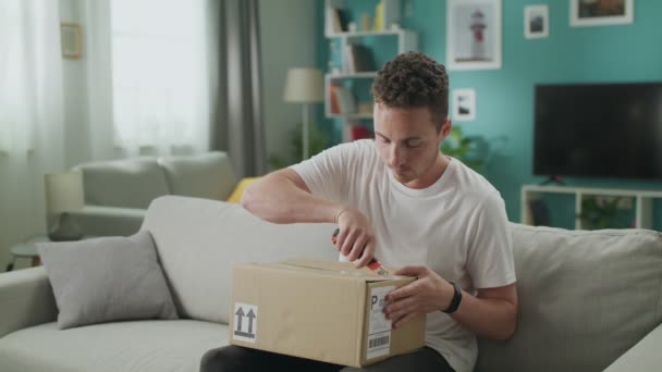 Jongeman zit op de bank in zijn woonkamer en opent kartonnen doos pakket — Stockvideo