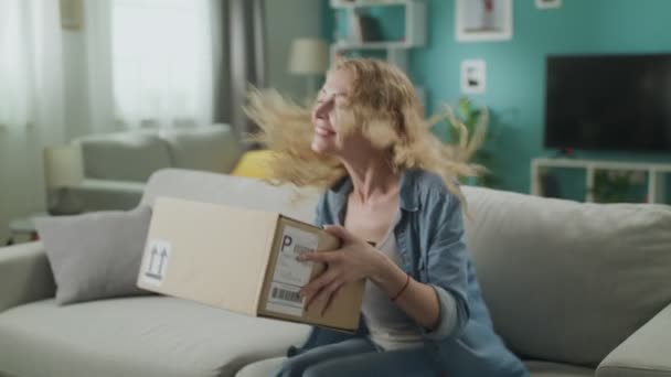 Dolly in van jonge vrouw opent een kartonnen perceel op de Bank in zijn woonkamer — Stockvideo