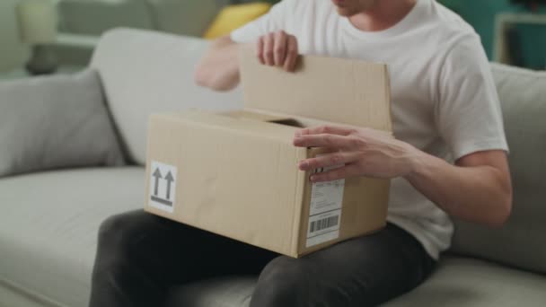 Dolly out of Young Man opent een kartonnen pakje op de Bank in zijn woonkamer — Stockvideo