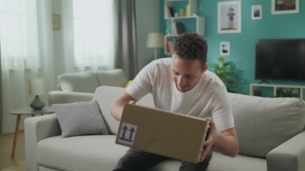 Young man enters his living room with cardboard box package, starts opening it — Stock Video