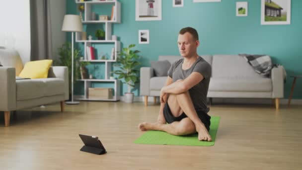 Un joven estudia yoga siguiendo las instrucciones de una tableta — Vídeos de Stock