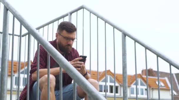 L'uomo è seduto con il telefono sulla scala antincendio — Video Stock