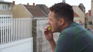 Büyük bir şehirde bir otelin balkonunda sandviç yiyen adam