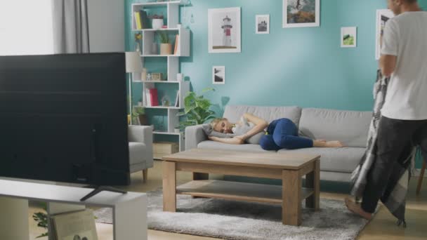 Joven cubre a una novia dormida con una manta en un sofá en la sala de estar — Vídeos de Stock