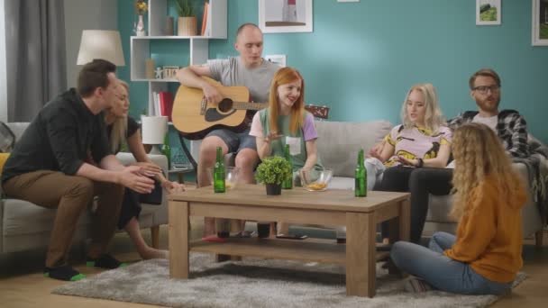Gruppe von Freunden singt Lieder, während ihr Freund im Wohnzimmer auf der Gitarre spielt — Stockvideo