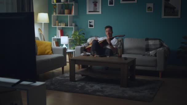 Osamělý mladý muž pije alkohol a hraje na kytaru doma v obývacím pokoji — Stock video