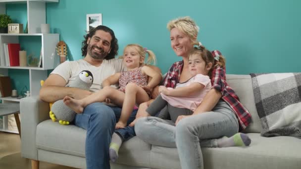 Familia feliz viendo la televisión. Pareja con hijas se sienta en un sofá gris y se ríe . — Vídeo de stock