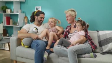 Kızları ile çift gri bir kanepede tv izlerken oturur ve gülüyor.