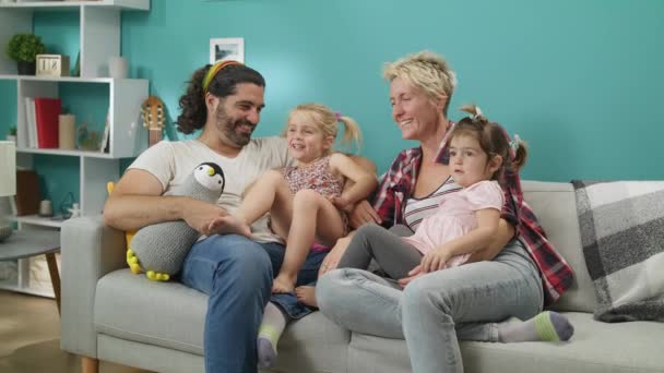 Paar mit Töchtern sitzt auf grauem Sofa vor dem Fernseher und lacht. — Stockvideo