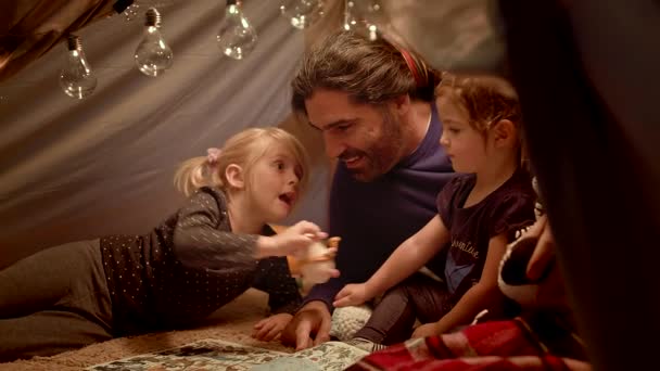 Father reads a book to his daughters in the evening in a toy tent in living room — Stock Video