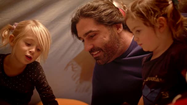 Vader leest 's avonds een boek naar zijn dochters in een speelgoed tent — Stockvideo