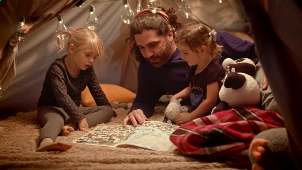 Папа с дочерьми читают книгу в вигваме ручной работы в комнате — стоковое видео