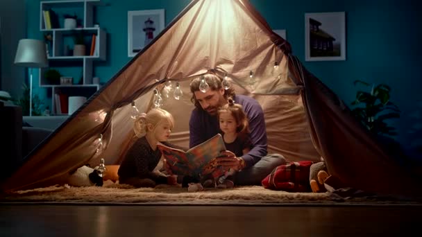 Papá feliz e hijas pequeñas leen un libro en una tienda de campaña en casa — Vídeo de stock