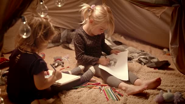 Hermanas dibujan con lápices en una carpa decorativa en la habitación por la noche — Vídeos de Stock