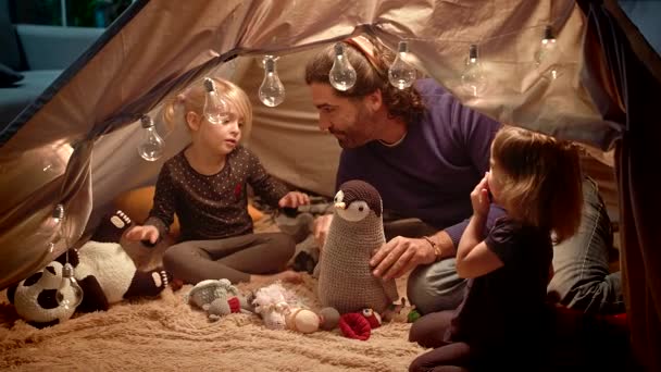 Papá con hijas juega con un pingüino de juguete en una tienda de campaña en casa . — Vídeos de Stock