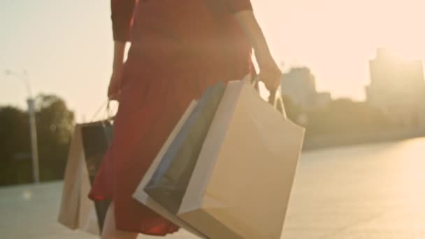 Femme en robe rouge avec des sacs à provisions marchant dans une ville au coucher du soleil. Mouvement lent — Video