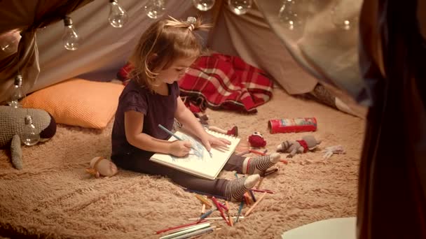 Menina desenha com lápis em uma cabana decorativa no quarto à noite — Vídeo de Stock