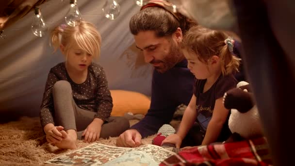 Padre feliz e hijas pequeñas leen un libro en una tienda en casa — Vídeos de Stock