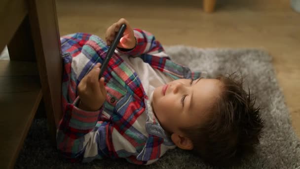 Küçük çocuk halı üzerinde yerde yatarken bir akıllı telefon üzerinde bir oyun oynar — Stok video