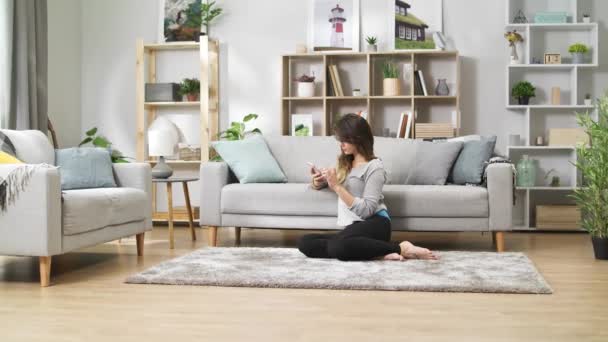 Una giovane ragazza con un telefono si siede sul pavimento in un accogliente soggiorno — Video Stock