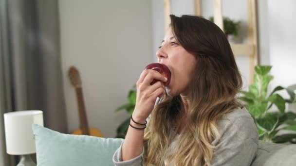 Молодая девушка ест яблоко, сидя на диване в гостиной — стоковое видео
