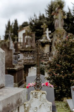 Gri, bulutlu bir günde mezar taşları ve taş mezarlıklarla dolu bir mezarlığın ön planında süslü paslı bir demir haç..