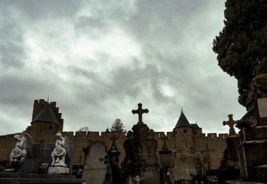 Arka planda Carcassonne kalesinin duvarları ve bulutlu gökyüzünün altında melek heykelleri ve taş haçlarla dolu bir ortaçağ mezarlığı..