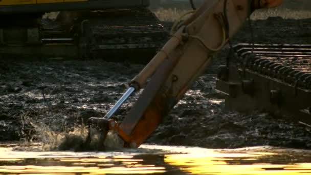 Cubo de la excavadora cava el suelo desde el fondo del río al atardecer. Limpieza y profundización del canal . — Vídeo de stock