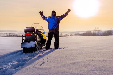 Snowmobile adventure. Man standing on snowy mountain in winner pose with raised hands enjoying view and achievement on sunset winter day. Freedom concept. clipart