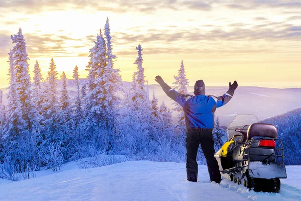 Man standing on snowy mountain near snowmobile enjoying view and achievement on sunset winter day.