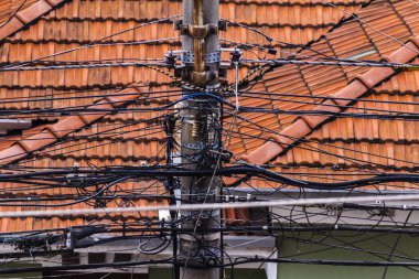 Elektrik direğine dolanmış kablolar kırmızı kiremit çatısına karşı karmaşık bir ağ oluşturur. Kentsel elektrik altyapısı zorluklarını gözler önüne seriyor.
