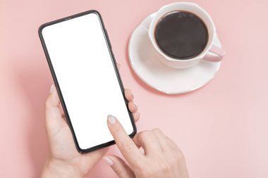 Taklit cep telefonu, akıllı telefon. Kadınların elleri pembe arka planda boş ekranlı bir cep telefonu tutuyor. Pembe masada bir fincan kahve.