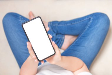 Görüntü görüntüsü boş beyaz ekran cep telefonu. Kadın elleri evdeki koltukta cep telefonuyla mesajlaşıyor. Reklam metni için boş alan. Kot pantolonlu genç bir kadın akıllı telefon ekranına bakıyor. Evdeki kanepede oturuyor. Üst görünüm