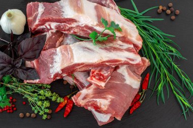 Çiğ domuz kaburgası ve baharatlar siyah bir taş kesme tahtasına serilmiş. Koyu siyah arka planda taze otlu çiğ domuz pirzolası. Çiğ domuz pirzolası, biber, fesleğen, sarımsak ve sos. Yakın plan.